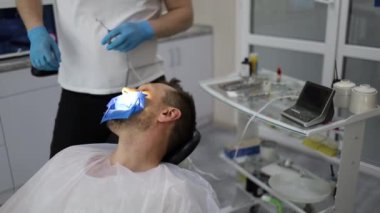 Bir erkek dişçi, sandalyede yatay olarak yatan bir adamın dişinde delik açma sürecinde. Mavi üniformalı ve maskeli ortodontist.