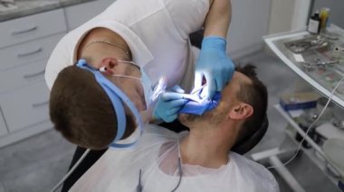 Bir erkek dişçi, sandalyede yatay olarak yatan bir adamın dişinde delik açma sürecinde. Mavi üniformalı ve maskeli ortodontist.