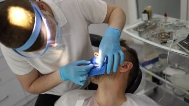 Bir erkek dişçi, sandalyede yatay olarak yatan bir adamın dişinde delik açma sürecinde. Mavi üniformalı ve maskeli ortodontist.