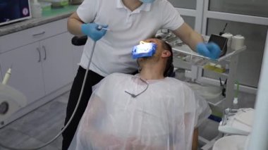 Bir erkek dişçi, sandalyede yatay olarak yatan bir adamın dişinde delik açma sürecinde. Mavi üniformalı ve maskeli ortodontist.