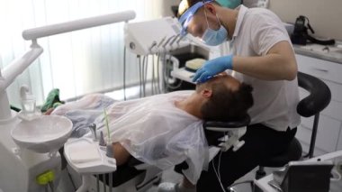 Bir erkek dişçi, sandalyede yatay olarak yatan bir adamın dişinde delik açma sürecinde. Mavi üniformalı ve maskeli ortodontist.