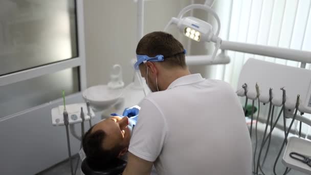 一个男牙医在一个水平躺在椅子上的男人的牙齿上钻一个洞 那个穿着蓝色制服和面具的牙齿矫正师 — 图库视频影像