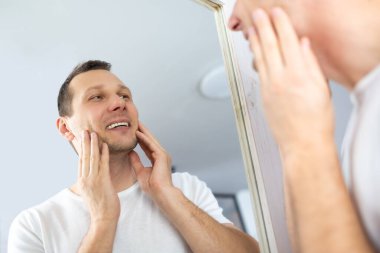 Caucasian man touching face applying moisturizer on face, standing near mirror in modern bathroom. Male facial skincare routine