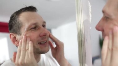 Caucasian man touching face applying moisturizer on face, standing near mirror in modern bathroom. Male facial skincare routine