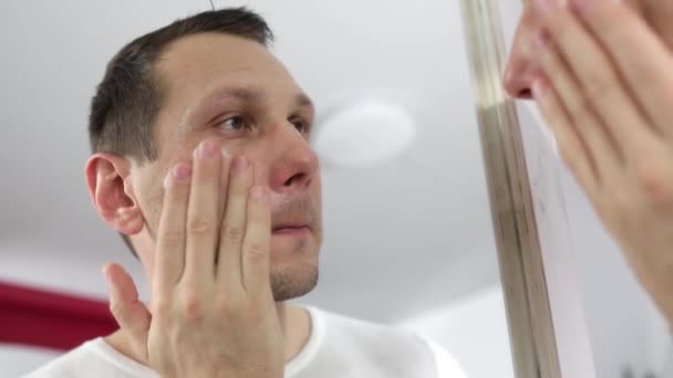 顔に保湿剤を適用白人男性のタッチ顔は 現代のバスルームでミラーの近くに立っている 男性の顔のスキンケアルーチン — ストック動画