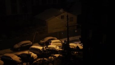 Falling snow in residential area at night. City courtyard night view in blizzard. Night town street with burning street lamps and snow covered parked cars during snowing