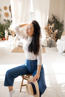 Cozy homely atmosphere. Stylish Asian woman in jeans sits on a chair over a bed and house plants. Soft colors.