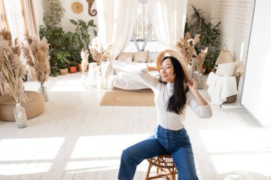 Cozy homely atmosphere. Stylish Asian woman in jeans sits on a chair over a bed and house plants. Soft colors.