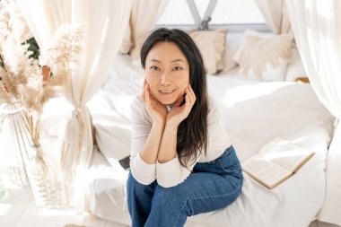 Young beautiful middle aged Asian woman sitting on bed at home with natural light.