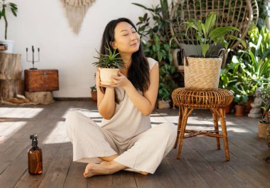 A beautiful Asian woman takes care of houseplants. A woman watering ficus in a red pot. Lots of plants in a wooden house. Concept: home gardening and floriculture.
