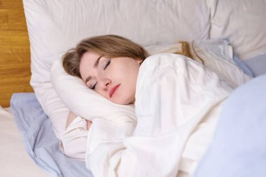 Calm serene beautiful young lady wears pajamas, lying down in a dream, relaxing sleep in a cozy white bed on a soft pillow covered with a blanket, enjoying a good healthy sleep concept, top view