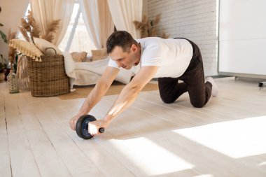 Athletic man is doing exercises with an abs wheel in living room at home. Sport and fitness. Workout and wellness concept. Fitness workout of guy. High-intensity interval training.