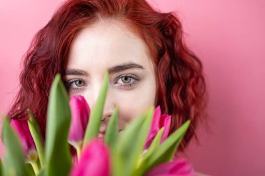 Güzel, kızıl saçlı bir kızın portresi. Elinde bir buket süsen çiçeği ve pembe arka planda izole edilmiş laleler var.