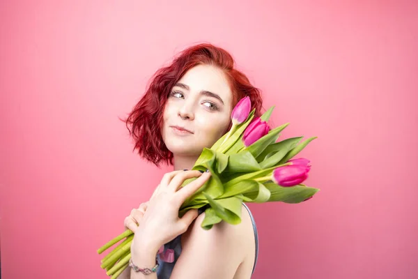一个漂亮的红头发少女拿着一束带粉色背景的虹膜和郁金香的画像 — 图库照片
