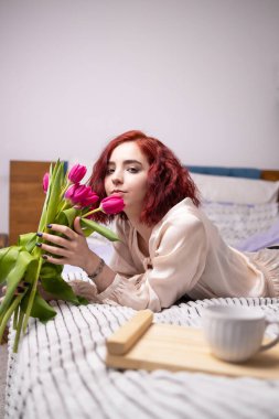 Genç kızıl saçlı kız elinde bir kupa sıcak kahve tutuyor. Yatakta lalelerle bahar kahvaltısı.