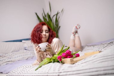 Beyaz gecelikli güzel kadın otelde yatakta kahvaltı yapıyor. Laleler, bir fincan kahve ahşap tepside servis edilir. Güzel kadın mutlu ve güneşli sabahın tadını çıkarıyor.