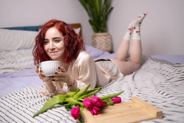 Young happy red head girl holding a mug of hot coffee in hands. spring breakfast with tulips on bed.