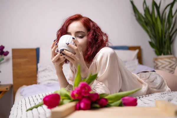 Young red head girl holding a mug of hot coffee in hands. spring breakfast with tulips on bed.