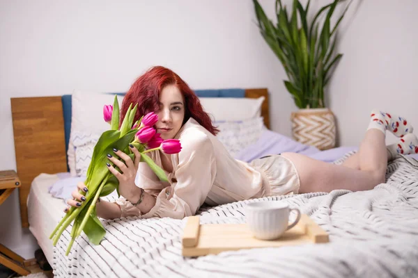 Young red head girl holding pink tulips in hands. spring breakfast with tulips on bed.