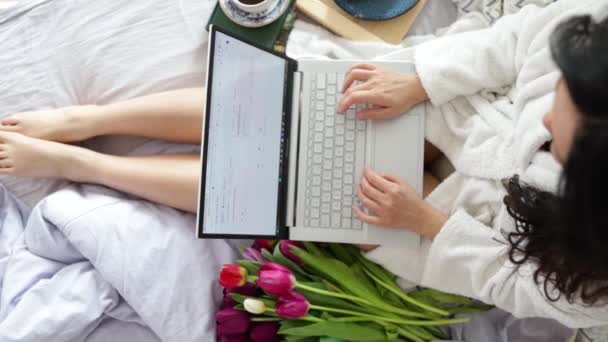 午前中にベッドの上に座っている間 ノートパソコンのブログとパジャマで肯定的な中年の女性 最上階だ 自宅で余暇を過ごしながら ラップトップで入力陽気な長い髪の女性 — ストック動画
