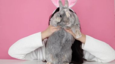 Tavşan kulaklı komik mutlu kız pembe arka planda gri tavşanı tutuyor.