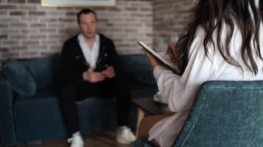 Kadın psikolog not alıyor ve müvekkilini psikoterapi seansında dinliyor. Kanepede oturup terapistle konuştuktan sonra zihinsel sağlık sorunları olan odaklanmamış yetişkin bir adam.