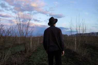 Şapkalı gizemli bir adamın arka planı kuru otların arasında yürüyor. Bulutlu karanlık gökyüzü.