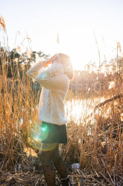 Beyaz yün kazaklı zarif genç kadın, sonbahar parkında sazlıkların arkasında poz veriyor. Sonbahar modası ve güzelliği.