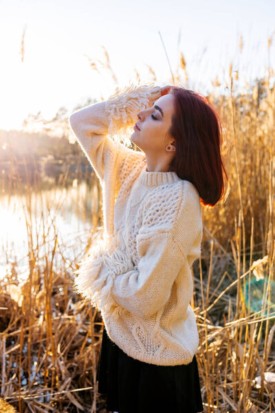 Elegant young woman in a white woolen sweater poses in the autumn park on the background of reeds. Autumn fashion and beauty.