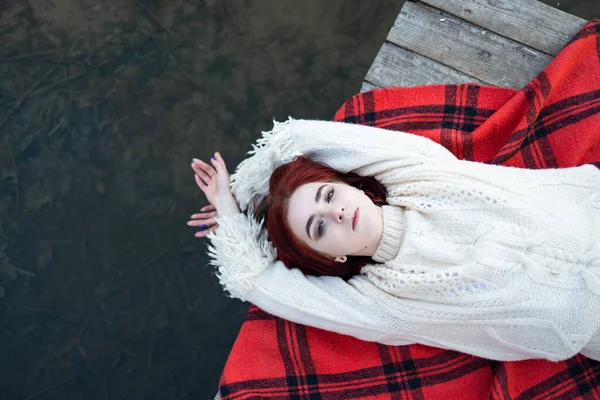 穿着毛衣的红头发姑娘躺在自然湖畔的一个码头上 — 图库照片