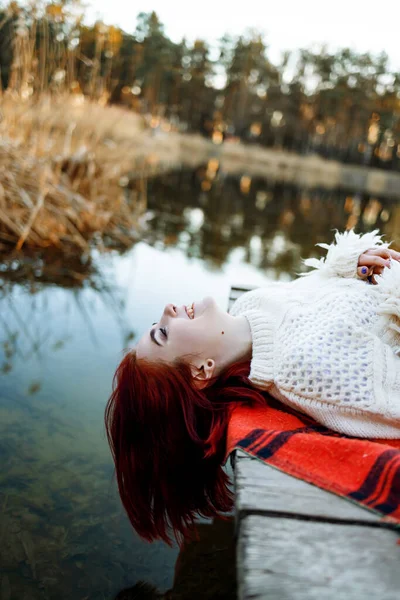 穿着毛衣的红头发姑娘躺在自然湖畔的一个码头上 — 图库照片