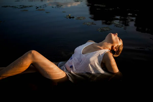 Woman in dress floating in water of pond with seaweed. Water overflowing her body when she floating on the surface.