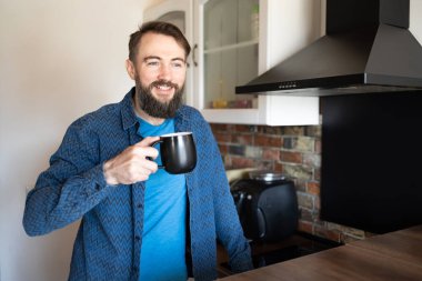 Yakışıklı, iyimser, mutlu, çekici bir adamın elinde taze kahveyle mutfaktaki pencereye baktığı bir fotoğraf.