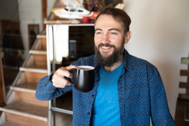 Yakışıklı, iyimser, mutlu, çekici bir adamın elinde taze kahveyle mutfaktaki pencereye baktığı bir fotoğraf.