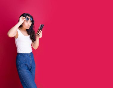Yazlık kıyafetli Asyalı kadın heyecanla akıllı telefona bakıyor, kırmızı stüdyo arka planında güneş gözlüğünü kaldırıyor. Şaşkın kadın internette yaz indirimini öğreniyor..