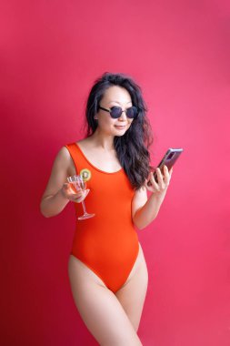 Kırmızı mayo giyen Asya kökenli genç bir kadın kırmızı arka planda cep telefonunu kapalı tutuyor. Yaz tatili deniz tenli güneş kavramı.