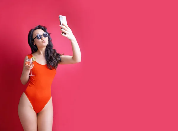 身穿红色泳衣的年轻亚洲女人拿着鸡尾酒 用红色背景的手机自拍 暑假海上休息日光浴的概念 — 图库照片