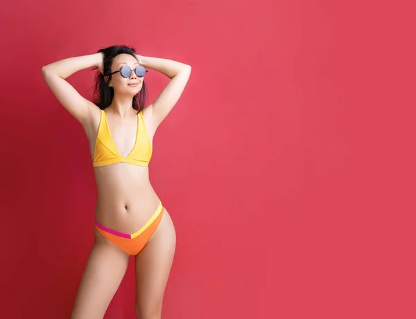 stock image fashion portrait of young beautiful asian woman in yellow swimwear. Carefree model with perfect body. Hot tanned female isolated on red in studio in summer swimsuit.