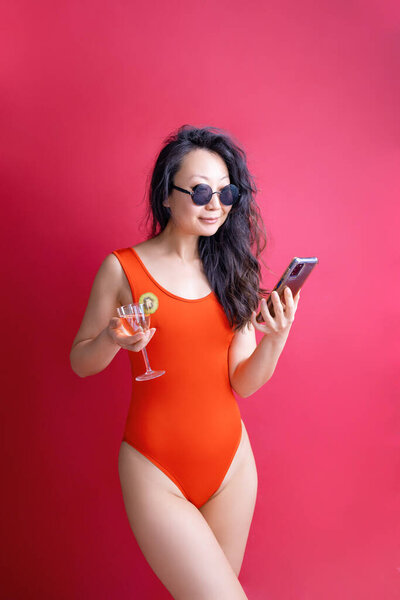 Young woman of Asian ethnicity in red swimsuit hold cocktail mobile phone isolated on red background. Summer vacation sea rest sun tan concept.