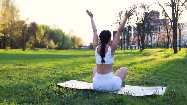 在城市公园里 一个无法辨认的身材苗条的年轻女子以莲花的姿势坐在瑜伽垫上 双手高举 晴天女性户外运动瑜伽的背景图 — 图库视频影像