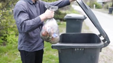 Siyah çevre dostu çöp torbasını büyük plastik yeşil çöp konteynırına atan adam. Çöpü dışarı çıkar.