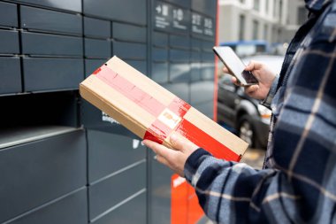 Posta terminalinin yanında elinde kutu olan bir adam. Paket teslimat makinesi. Elinde bir karton kutu tutan kişi. Posta ve posta servisi, online alışveriş, e-ticaret konsepti