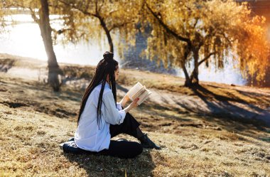 Rasta saçlı genç bir kadın bahar parkında kitap okuyor..