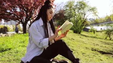 Rasta saçlı genç bir kadın bahar parkında kitap okuyor..