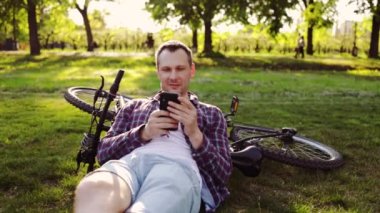 Yaz parkında bisikletin yanında çimlerde yatan genç adam. Bisiklet sürdükten sonra dinlenen ve cep telefonu kullanan mutlu kişi.