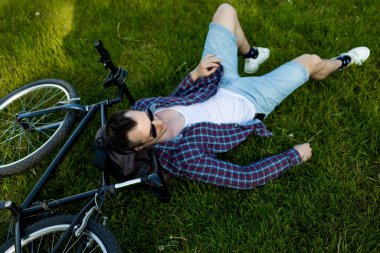 Yaz parkında bisikletin yanında dinlenen genç adam. Bisiklet sürdükten sonra mutlu insan, gözleri kapalı, doğada sessizliğin tadını çıkarıyor. Spor, açık hava aktivitesi, fitness konsepti