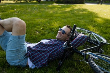 Yaz parkında bisikletin yanında dinlenen genç adam. Bisiklet sürdükten sonra mutlu insan, gözleri kapalı, doğada sessizliğin tadını çıkarıyor. Spor, açık hava aktivitesi, fitness konsepti