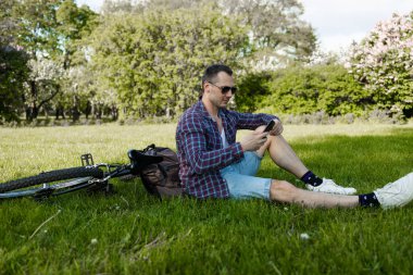 Adam telefonu Park'taki günbatımında kullanarak bisikleti ile. Çimenlerin üzerinde oturan ve onun Smartphone'da yazarak Beyaz delikanlı. Yaşam tarzı ve seyahat kavramları