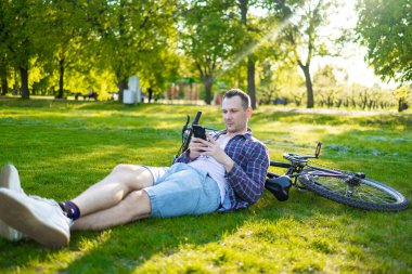Adam telefonu Park'taki günbatımında kullanarak bisikleti ile. Çimenlerin üzerinde oturan ve onun Smartphone'da yazarak Beyaz delikanlı. Yaşam tarzı ve seyahat kavramları
