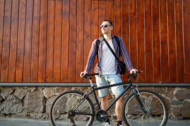 Sokak tarzı giyinmiş, bisikleti duvara dayalı duran ve başka tarafa bakan mutlu, beyaz bir adam.